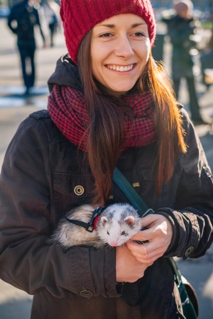 Smiling young woman with her pet ferret in hands walking together on a street on a sunny autumn day. Woman and a pet concept.の写真素材