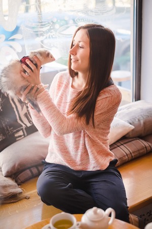 Beautiful girl with brown hair having fun with her pet ferret at a cafe window sill on a sunny cold morning. Woman and a pet concept.の写真素材