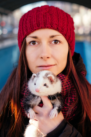 Closeup portrait of the young woman in red cap and her pet ferret in foreground on a sunny cold day in the city. Woman and a pet concept.の写真素材