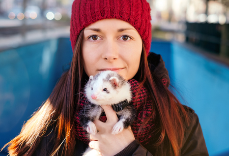 Closeup of the young woman in red cap and her pet ferret in foreground on a sunny cold day in the city. Woman and a pet concept.の写真素材
