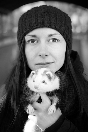 Black and white. Closeup portrait of the young woman with long hair and in a cap standing with her pet ferret in foreground on a sunny cold day in the city. Woman and a pet concept.の写真素材