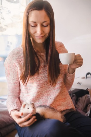 Young woman having her hot drink in a cafe while her pet ferret resting on her legs. Woman and a pet concept.の写真素材