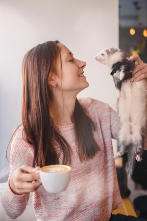 Portrait of happy girl with a cup of cappuccino in her hand and her pet ferret in the other looking and leaning to each other in a cafe.Woman and a pet concept.の写真素材
