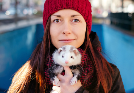 Closeup of the young woman in red cap and her pet ferret in foreground on a sunny cold day in the city. Looking at camera. Woman and a pet concept.の写真素材