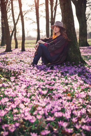 Beautiful red head lady leaning against tree on a warm, sunny morning in the Spring forest with a lilac crocus carpet instead of grass. Vertical orientation. Inspiration and wanderlust travel concept.の写真素材