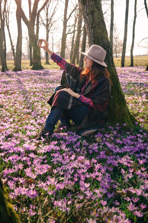 Beautiful red head girl in a hat and colorful poncho, matching lilac crocuses around, taking selfie of Spring blossoming beauty. Vertical orientation. Inspiration and wanderlust travel concept.の写真素材