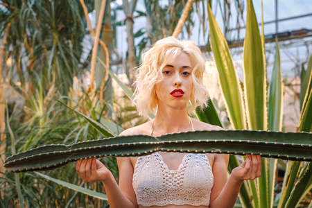 Closeup portrait of a beautiful female fashion model with big dreamy eyes, full red lips wearing sexy crochet top touching cactus with both hands at the greenery.の写真素材