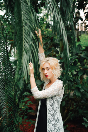 Beautiful young fashion model with curly blonde hair wearing black dress and white lace cardigan posing next to the palm, reaching for leaves at the tropical garden, looking at camera.の写真素材