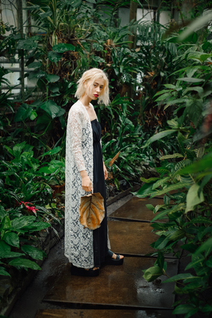 Full length portrait of young female model wearing white lace cardigan and black dress standing on a path among tropical plants at greenhouse. Holding big dry leaf in hand, looking at camera.の写真素材