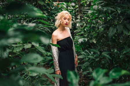 Fashion portrait of a sensual female model standing in a black dress and white lace cardigan posing with one bear shoulder, looking dreamy, distant in a rainforest, surrounded by tropical plants.の写真素材
