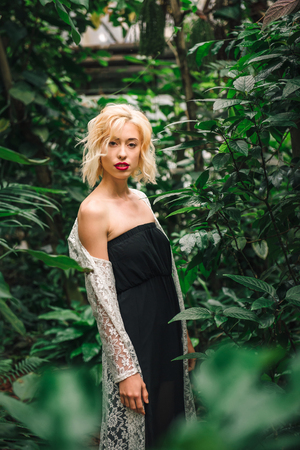 Fashion portrait of a young perky female model wearing black dress and a white lace cardigan posing with a bear shoulder in a rainforest, looking at camera.の写真素材