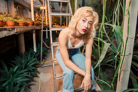 Beautiful female model with short curly hair wearing jeans and top with bear shoulders sitting on a stepladder surrounded by succulents. Looking at camera at botanical garden.の写真素材
