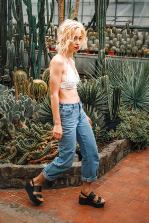 Full length portrait of slender perky female model walking along cacti and other succulents at botanical garden, wearing white top and jeans.の写真素材