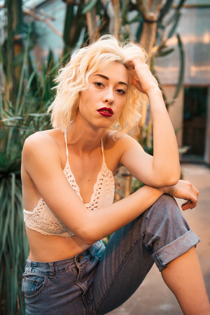 Fashion portrait of the tempting young woman with curly blonde hair and seductive red lips posing in greenhouse. Wearing sexy top and jeans sitting with one leg bent at the knee and one arm in her hair. Looking at camera.の写真素材