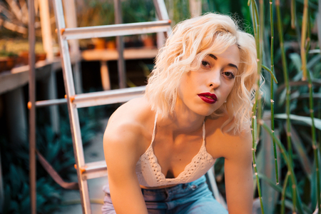 Closeup fashion portrait of a young sensual blonde model with penetrative look, wearing sexy knitted white top and jeans, sitting on a stepladder at greenhouse. Looking at camera.の写真素材
