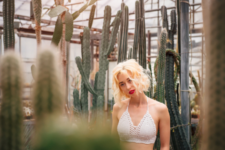 Young and slender female fashion model in a sexy white crochet top standing among cacti looking dreamy and far away at a greenery.の写真素材