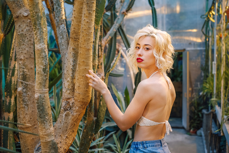 Portrait of an attractive young model in a sexy top showing bear skin and high waist jeans standing close to the thorny trunk of a tropical plant at the greenery.の写真素材