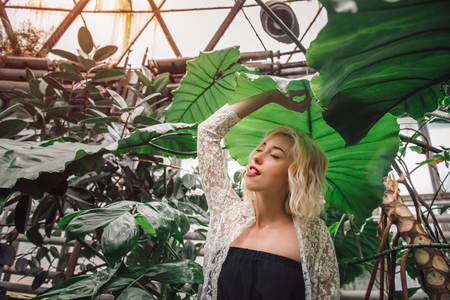 Fashion portrait of attractive female model standing under a giant leaf with eyes closed playfully sticking tongue out, holding that same leaf with one hand at the botanical garden.の写真素材