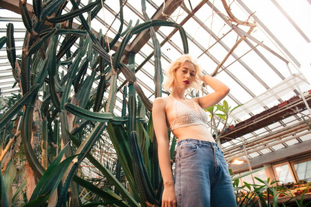 Beautiful young woman posing with one arm in her curly blonde hair at tropical botanical garden among cacti. Looking at camera. Low angle view.の写真素材