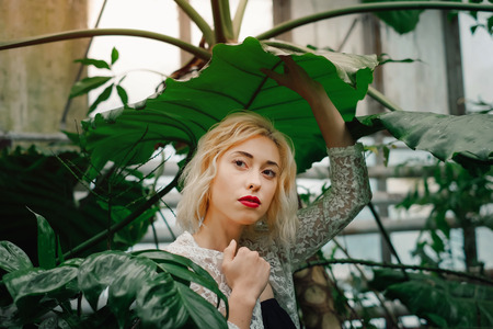 Portrait of dreamy fashion model standing under the giant leaf of exotic plant at greenery. Holding the leaf with one hand, wearing white lace cardigan.の写真素材