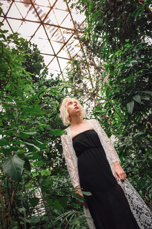 Portrait of an attractive female model with a dreamy look wearing black dress and white lace cardigan standing among tropical plants in a botanical garden on a background of its glass roof.Low angle view.の写真素材