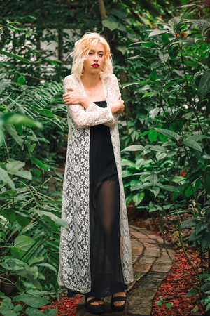 Full length portrait of a slender young female model in a long black dress and white lace cardigan standing with arms crosses on a brick path at a tropical garden, looking at camera.の写真素材