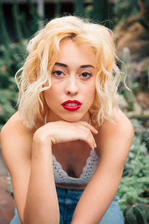 Closeup portrait of a beautiful young female model with curly blonde hair, big dreamy eyes and full red lips wearing knitted top and jeans sitting among tropical plants . Looking at cameraの写真素材
