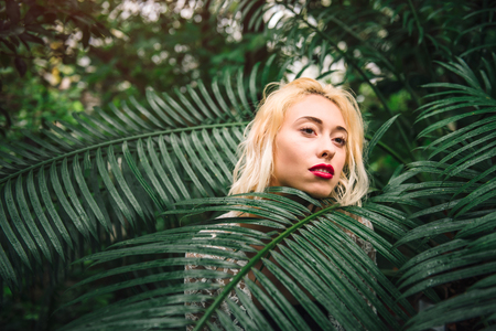 Closeup portrait of sensual female fashion model with damp blond hair and red lips standing behind the palm branch, covered in it. Having a dreamy look, at the botanical garden.の写真素材