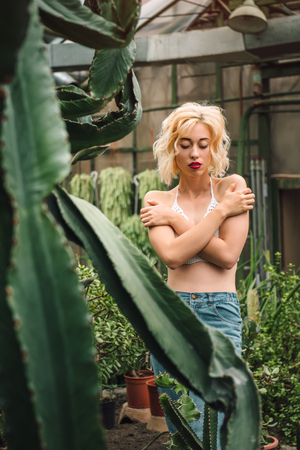 Sensual slim female model wearing top and jeans standing humble with her arms crossed with eyes closed at the greenery full of tropical plants.の写真素材