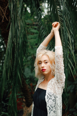 Portrait of beautiful sensual female model in a black dress and white lace cardigan posing with raised arms leaning against palm leaves at greenery. Looking at garden.の写真素材