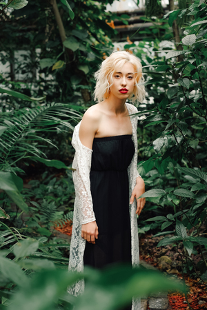 Portrait of beautiful slender female model with perfect curly blonde hair wearing black dress and white lace cardigan revealing one shoulder. Looking at camera, standing in a tropical rainforest.の写真素材