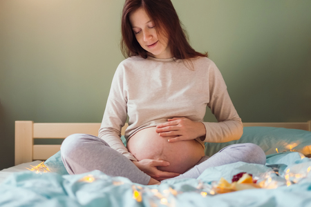 Portrait of beautiful young caucasian woman with messy hair in pajamas sitting on bed, caressing her belly with both arms at bedroom in the morning. Pregnancy and expecting concept.の写真素材