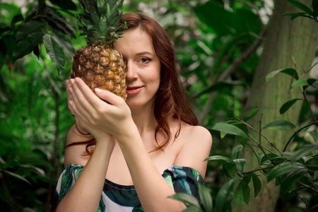 Beautiful young caucasian woman with long red hair and bare shoulders, holding a pineapple in her hands high up,hiding half of a face at tropical forest. Looking at camera.の写真素材