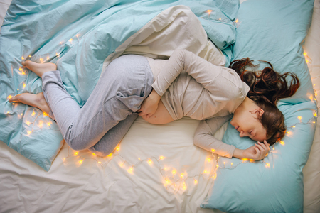 Young caucasian woman expecting baby sleeping on a bed decorated with lights,expressing upcoming fest and miracle of life, wearing comfortable home clothes. Top view. Pregnancy and expecting concept.の写真素材