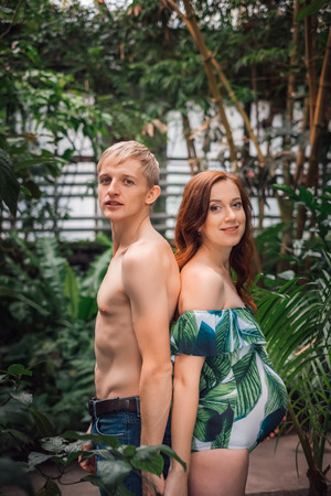 Caucasian couple of loving young parents expecting baby standing back to back, topless blonde daddy in jeans, and redhead mom in a floral swimsuit at tropical forest. Pregnancy and expecting concept.の写真素材
