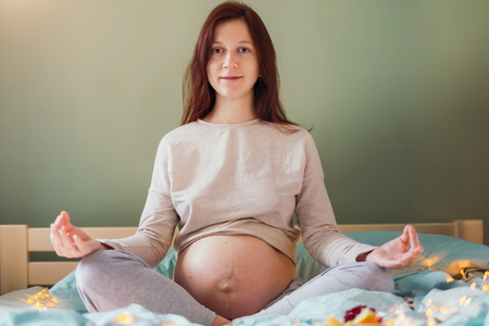 Portrait of beautiful young caucasian woman with messy hair in pajamas meditating on a bed, doing mudras at bedroom in the morning. Pregnancy and expecting concept.の写真素材
