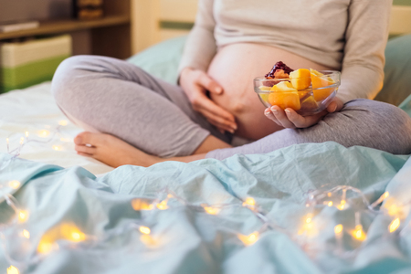 Belly of pregnant caucasian woman sitting on bed, holding a bowl with fruit salad in one hand, and supporting her belly with other. At bedroom in the morning.Healthy pregnancy diet concept.の写真素材