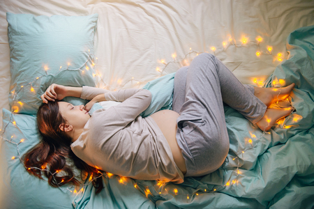 Young caucasian woman expecting baby sleeping on a bed decorated with lights,expressing upcoming fest and miracle of life, wearing comfortable home clothes. Top view. Pregnancy and expecting concept.の写真素材