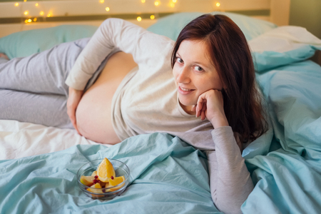 Young caucasian pregnant woman in comfortable home clothes lying on a bed next to the bowl of fruit salad, leaning on one hand and touching her bellyの写真素材