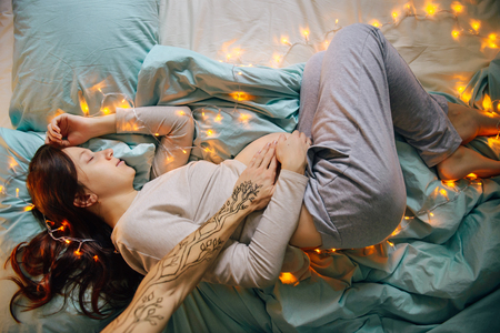Young caucasian woman expecting baby sleeping on a bed decorated with lights,expressing upcoming fest and miracle of life, wearing comfortable home clothes. Top view. Pregnancy and expecting concept.の写真素材
