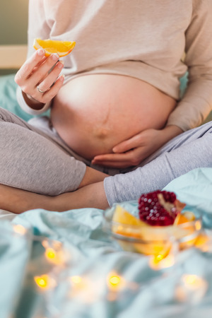 Belly of pregnant caucasian woman sitting on bed, holding a slice of orange in one hand, and supporting her belly with other, bowl with fruits. Healthy pregnancy diet concept.の写真素材