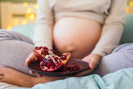 Closeup portrait of pregnant caucasian woman in pajamas holding plate of pomegranate in front of her belly, sitting on bed on xmas lights' background in the morning.Healthy pregnancy diet concept.の写真素材