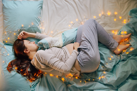 Young caucasian woman expecting baby sleeping on a bed decorated with lights,expressing upcoming fest and miracle of life, wearing comfortable home clothes. Top view. Pregnancy and expecting concept.の写真素材