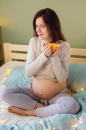 Portrait of pregnant caucasian woman with messy hair in pajamas sitting on bed holding a bowl of fruits in her hands at bedroom in the morning.Looking at camera.Healthy pregnancy diet concept.の写真素材