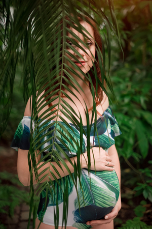 Portrait of attractive feminine woman expecting baby wearing floral swimsuit, standing behind palm leaf, holding belly in her hands at botanical garden. Pregnancy and expecting concept.の写真素材