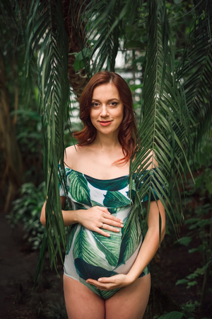 Attractive soon to be mom with a natural beauty and long red hair wearing floral swimsuit posing among palm leaves at tropical forest, caressing her belly with hands, looking happy and full of life.の写真素材