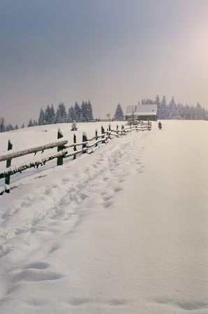 Remote traditional cottage in mountains after snow storm and freshly beaten track along the wooden fence. Shot on film camera with noise and grainの写真素材