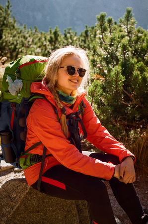 Happy blonde caucasian hiker in sunglasses and colorful outfit, lit with sun, resting on a stone, restoring her strength, surrounded by pine trees in High Tatras. Shot with film camera and grainの写真素材