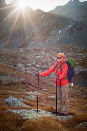 Full length portrait of happy young hiker in colorful outfit and sunglasses with walking poles posing at Popradske Pleso at sunset. Shot with film camera with grain.の写真素材