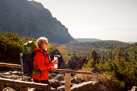 Blonde female backpacker in sunglasses and colorful clothes holding water flask in her hand while standing on a wooden bridge on a sunny day in High Tatras. Shot with film camera and grainの写真素材